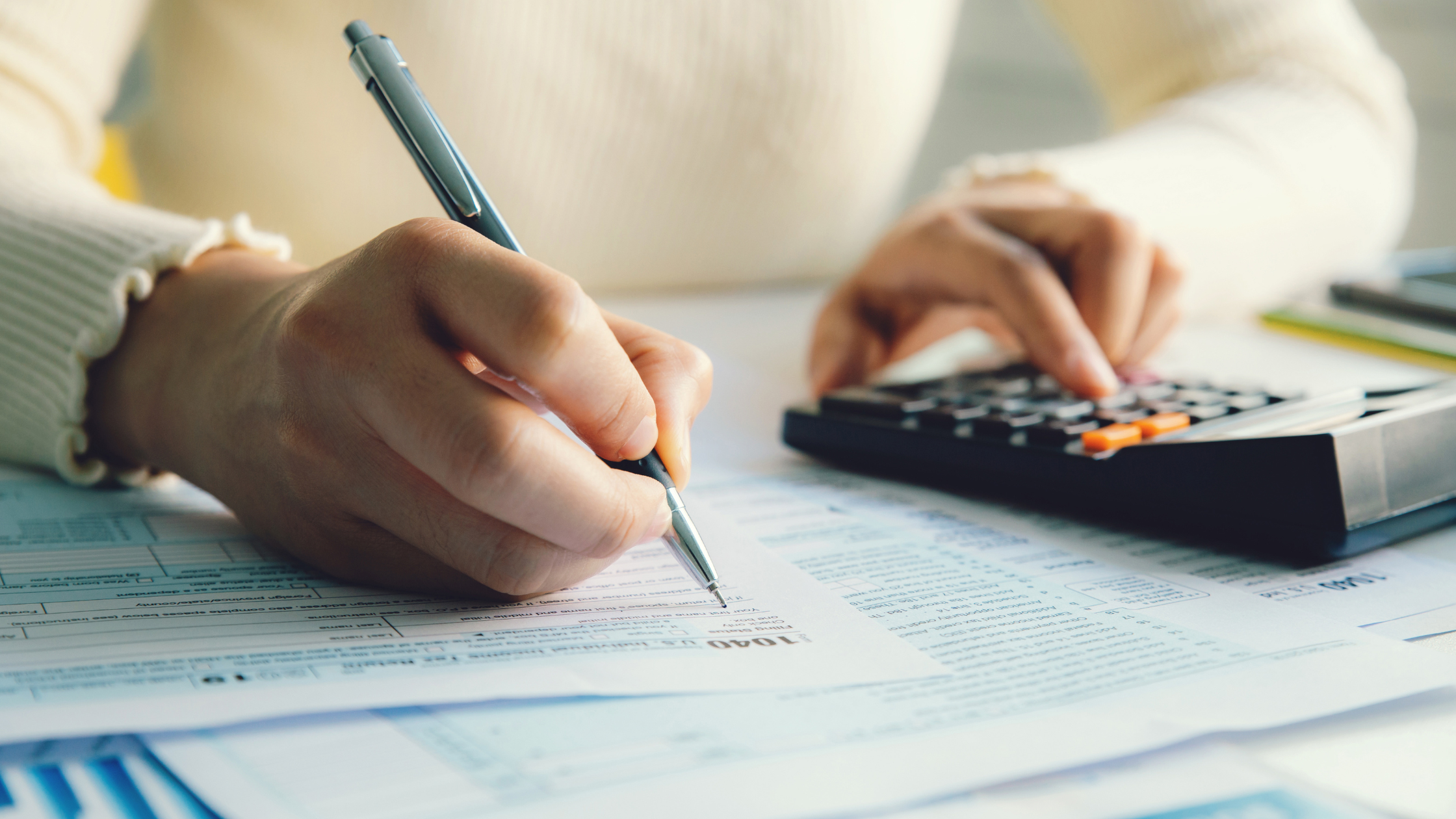 Close-up of a woman's hands holding a pen over tax papers with one hand and typing on a calculator with the other