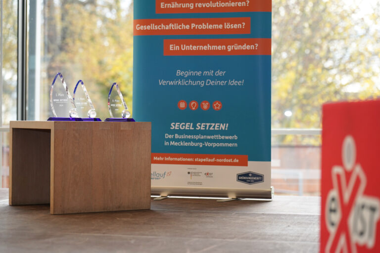 Ein Banner des Businessplanwettbewerbs Segel Setzen steht hinter dem Beistelltisch, auf dem drei Trophäen aus Glas stehen