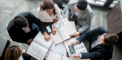 Symbolbild für Startups: Bild von oben aufgenommen: 5 Personen sitzen um einen Tisch, auf dem sich Arbeitsunterlagen befinden