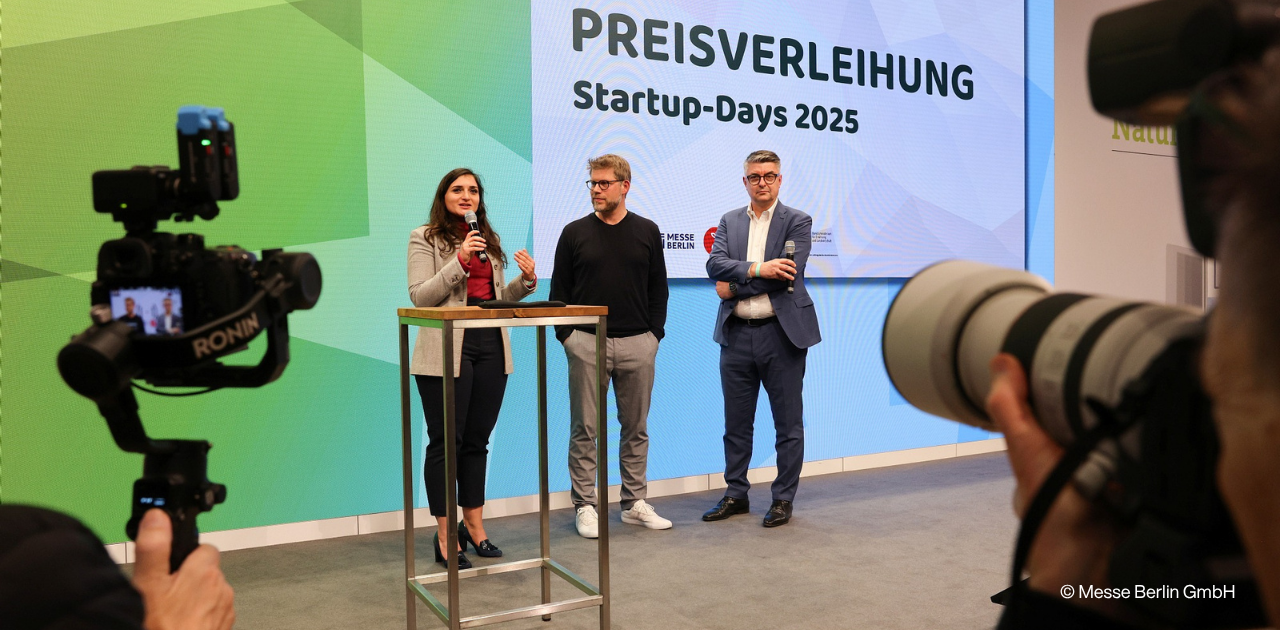 Drei Personen auf einer Bühne bei den Startup-Days 2025 in Berlin während einer Preisverleihung, mit zwei Kameras im Vordergrund.