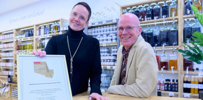 Wencke und Thomas Witte stehen in ihrer Feinkostboutique „Die Genusswelt“ in Schwerin. Wencke hält eine gerahmte Auszeichnung, während Thomas sich lächelnd auf den Tresen lehnt. Im Hintergrund sind Regale mit Gourmetprodukten wie Ölen und Essigen zu sehen.