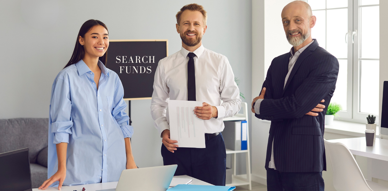 Drei Personen stehen in einem Büro vor einem Tisch mit Dokumenten und Laptop, im Hintergrund eine Tafel mit der Aufschrift „SEARCH FUNDS“.