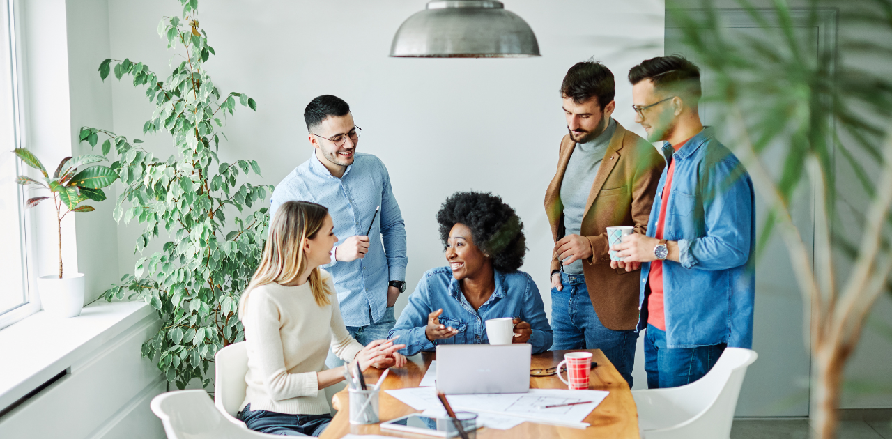 5 Personen verschiedener Ethnien stehen oder sitzen an einem Tisch und unterhalten sich