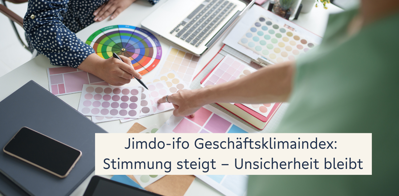 Arbeitsplatz mit Laptop, Farbfächern, Farbmusterkarten und Händen zweier Personen, die gemeinsam an Materialien arbeiten. Im Vordergrund befindet sich ein Textfeld mit der Aufschrift „Jimdo‑ifo Geschäftsklimaindex: Stimmung steigt – Unsicherheit bleibt“.