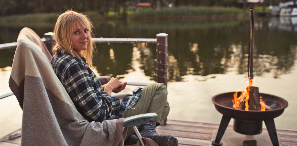 Anna‑Maria Polkowski, Gründerin von Locamii, sitzt auf einem Steg am Wasser neben einem Feuerkorb und notiert Ideen für die Weiterentwicklung der Erlebnisplattform.