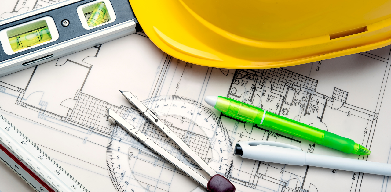 auf einem Tisch sind Bauarbeiterhelm, Stifte, Wasserwaage, Zirkel und Winkelmesser zu sehen, die auf einer Bauzeichnung liegen