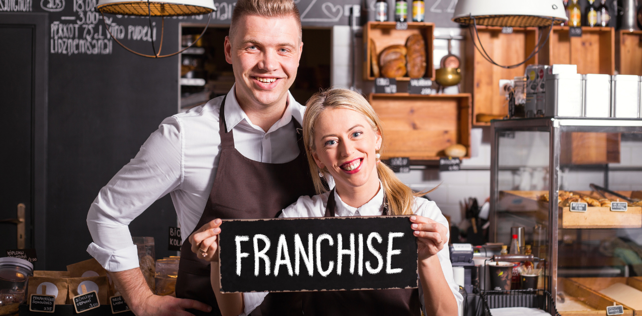Zwei Personen im Café halten ein Schild mit der Aufschrift „Franchise“ und stehen hinter einem Verkaufstresen mit Backwaren