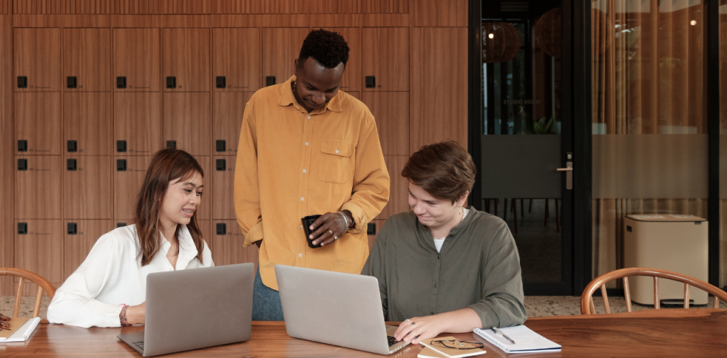 drei Personen sind an einem Tisch zu sehen und blicken auf einen von zwei Laptops