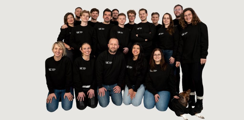 Gruppenfoto des Teams des Unisphere Innovation Campus (UIC) in schwarzen UIC-Pullovern vor hellem Hintergrund, mit einem Hund im Vordergrund.