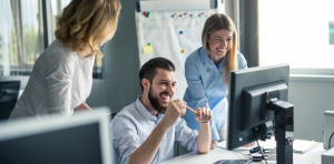 drei Personen sitzen bzw. stehen an einem Schreibtisch, blicken auf einen Monitor und lachen
