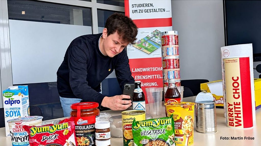 Auf einem Tisch liegen mehrere verpackte Lebensmittel. Lennart Standtke scannt mit einem Smartphone eine Verpackung.