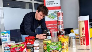 Auf einem Tisch liegen mehrere verpackte Lebensmittel. Lennart Standtke scannt mit einem Smartphone eine Verpackung.